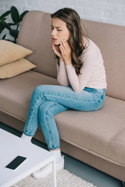 Vista ad alto angolo di giovane donna che soffre di dolore ai denti mentre è seduta sul divano a casa — Foto stock