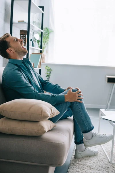 Seitenansicht eines Mannes, der auf der Couch sitzt und zu Hause unter Knieschmerzen leidet — Stockfoto