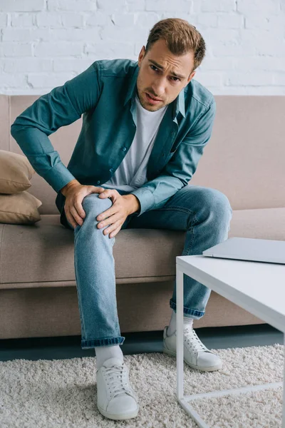 Mann leidet unter Knieschmerzen und blickt in Kamera, während er zu Hause auf dem Sofa sitzt — Stockfoto