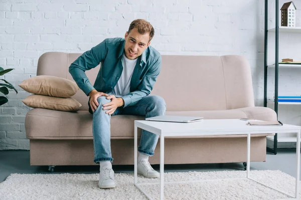 Young man suffering from knee pain and looking at camera while sitting on couch — Stock Photo