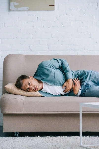 Young man lying on sofa and suffering from stomach ache at home — Stock Photo