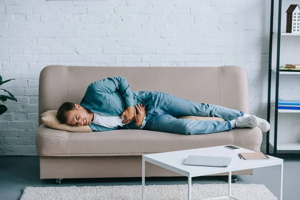 Young man lying on sofa and suffering from stomach pain at home — Stock Photo