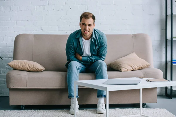 Young man with abdominal pain sitting on sofa and looking at camera — Stock Photo