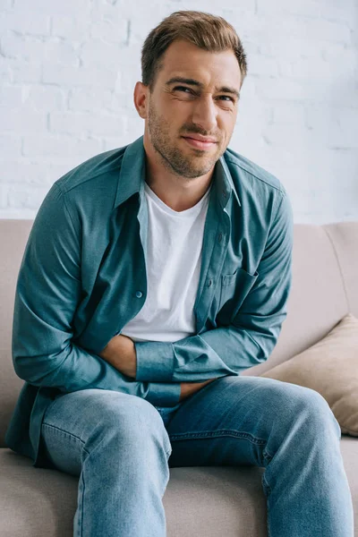 Young man with stomach ache sitting on sofa and looking at camera — Stock Photo