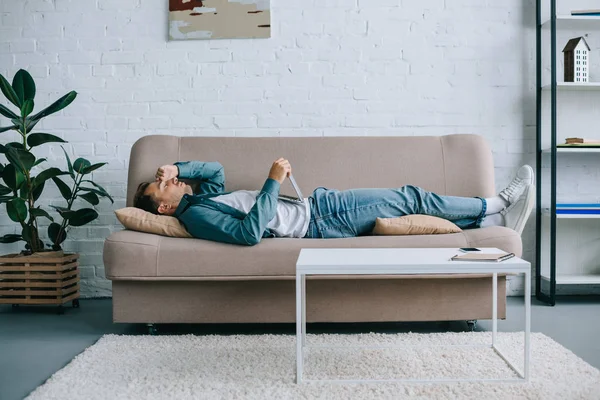 Боковой вид молодого человека, использующего ноутбук, страдающего головной болью на диване дома — стоковое фото