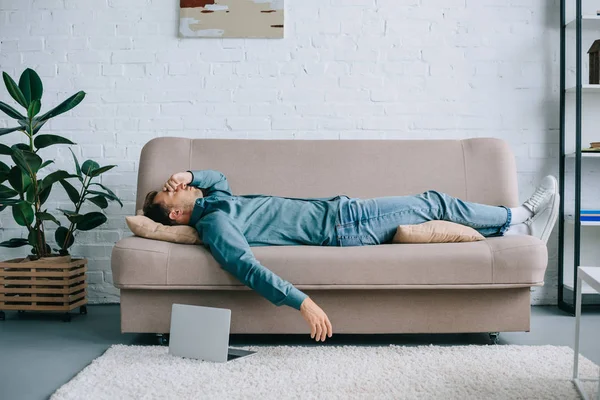 Young man with headache lying on sofa, laptop on carpet at home — Stock Photo