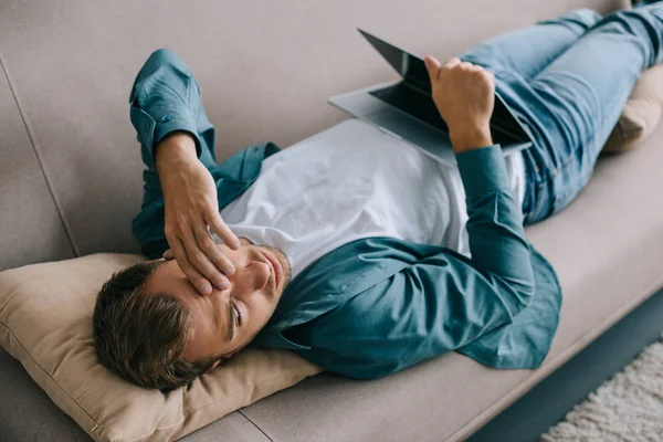 Joven con dolor de cabeza tumbado en el sofá y el uso de ordenador portátil - foto de stock