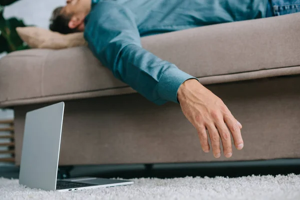 Vue rapprochée de l'ordinateur portable sur le tapis et l'homme malade couché sur le canapé — Photo de stock