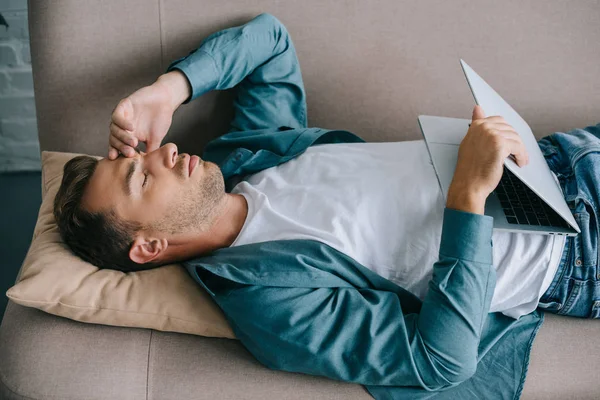 Jeune homme avec des maux de tête tenant ordinateur portable tout en étant couché sur le canapé — Photo de stock