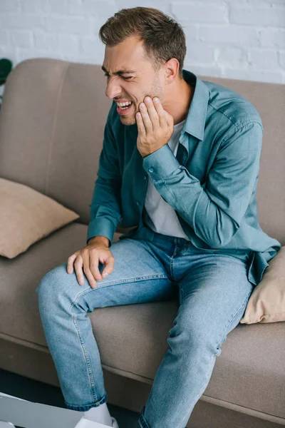 Молодой человек сидит на диване и страдает от зубной боли дома — стоковое фото