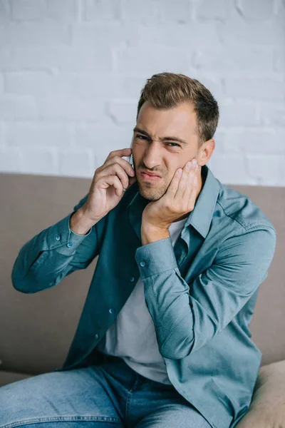 Giovane uomo che parla con lo smartphone e guarda la fotocamera mentre soffre di dolore ai denti a casa — Foto stock