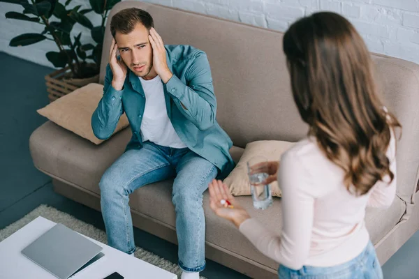 Joven con dolor de cabeza mirando a la mujer sosteniendo un vaso de agua y pastillas - foto de stock