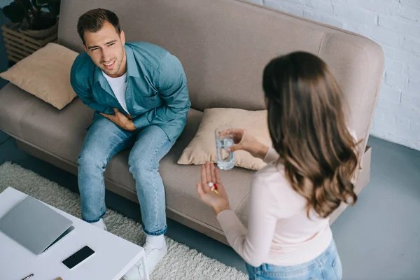 Vista de ángulo alto de la mujer que da vaso de agua y pastillas para el hombre con dolor de estómago - foto de stock