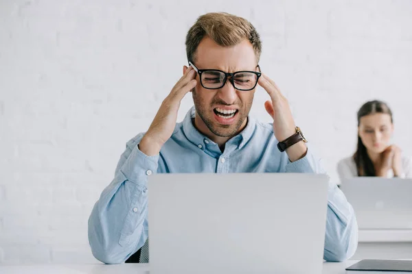 Joven hombre de negocios en gafas que sufren de dolor de cabeza mientras trabaja con el ordenador portátil en la oficina - foto de stock
