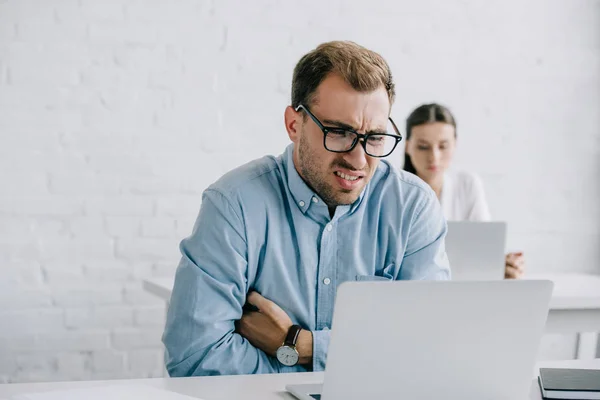 Young businessman in eyeglasses suffering from pain while working with laptop in office — Stock Photo