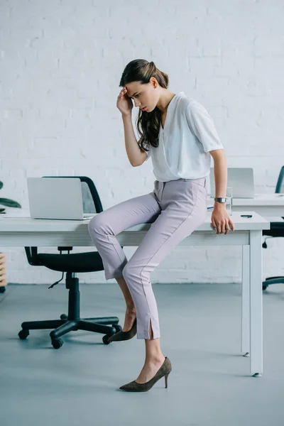 Молода бізнес-леді сидить на столі і страждає від головного болю в офісі — стокове фото