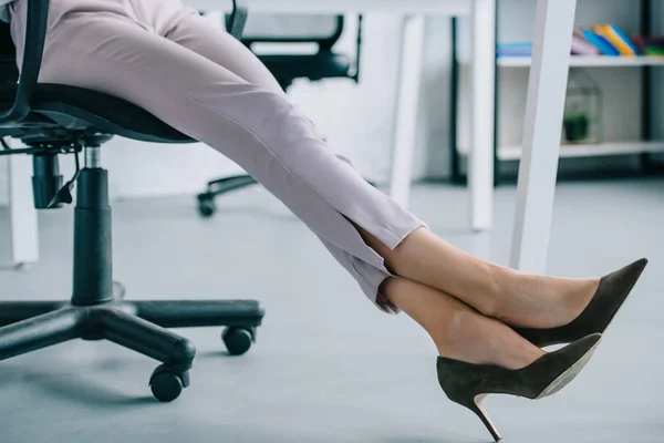 Tiro cortado de mulher de negócios em sapatos elegantes de salto alto sentado no local de trabalho — Fotografia de Stock