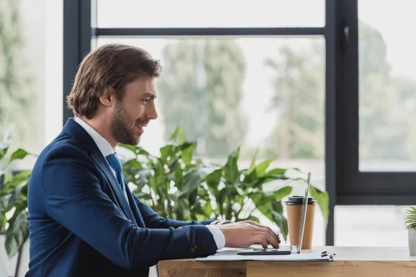 Vue latérale de sourire jeune homme d'affaires en utilisant un ordinateur portable dans le bureau — Photo de stock