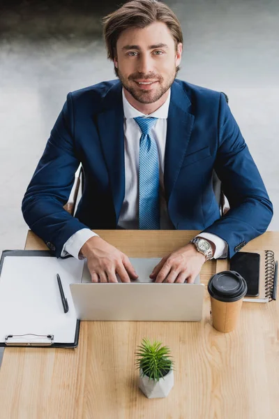 Vista de ángulo alto de joven hombre de negocios guapo usando el ordenador portátil y sonriendo a la cámara - foto de stock