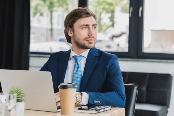 Beau jeune homme d'affaires utilisant un ordinateur portable et regardant loin dans le bureau — Photo de stock