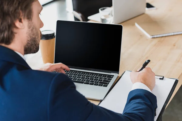 Recortado disparo de hombre de negocios tomando notas en el portapapeles y el uso de ordenador portátil con pantalla en blanco en el lugar de trabajo - foto de stock