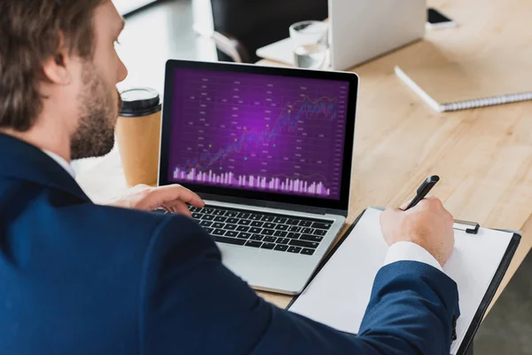 Recortado tiro de hombre de negocios tomando notas en el portapapeles y el uso de ordenador portátil con gráficos de negocios en la pantalla - foto de stock