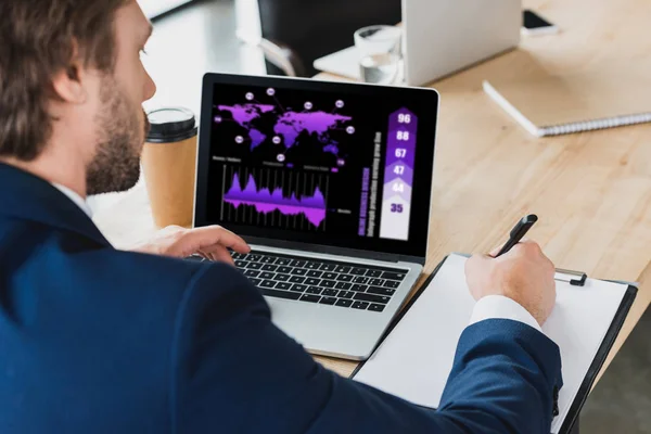 Cropped shot of businessman taking notes on clipboard and using laptop with business graphs on screen — Stock Photo