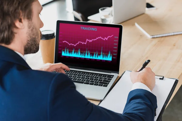 Recortado tiro de hombre de negocios tomando notas en el portapapeles y el uso de ordenador portátil con gráficos de comercio en la pantalla - foto de stock