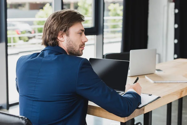 Back view of young businessman looking away while writing on clipboard and using laptop in office — Stock Photo