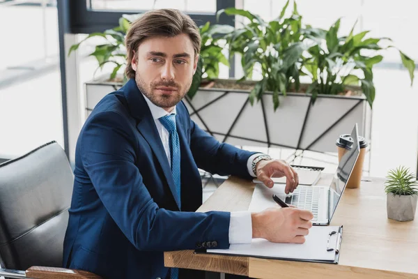 Handsome young businessman looking away while writing on clipboard and using laptop in office — Stock Photo