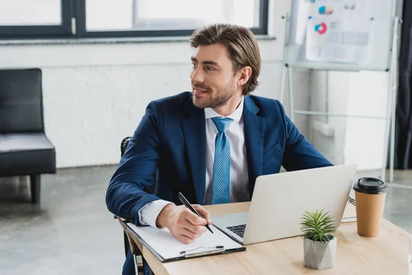 Lächelnder junger Geschäftsmann schaut weg, während er am Klemmbrett schreibt und Laptop im Büro benutzt — Stockfoto