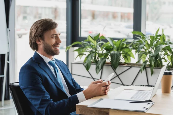 Side view of smiling young businessman using smartphone and laptop in office — Stock Photo