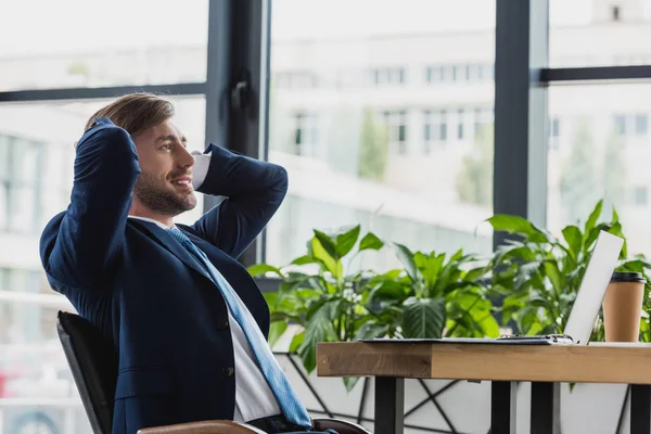 Sorridente jovem empresário sentado com as mãos atrás da cabeça e olhando para longe no escritório — Stock Photo