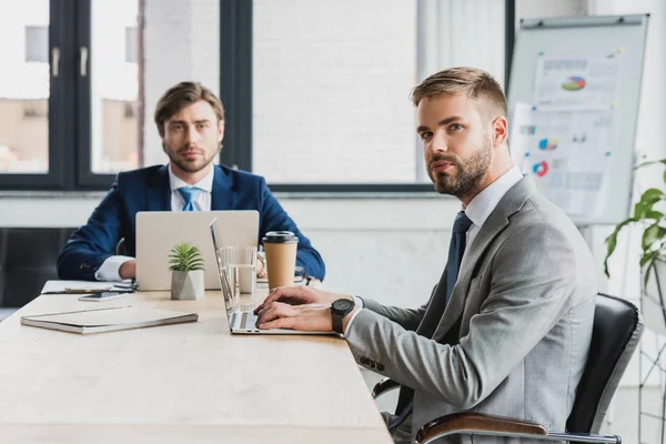 Jeunes hommes d'affaires en costumes à l'aide d'ordinateurs portables et en regardant la caméra tout en travaillant ensemble au bureau — Photo de stock