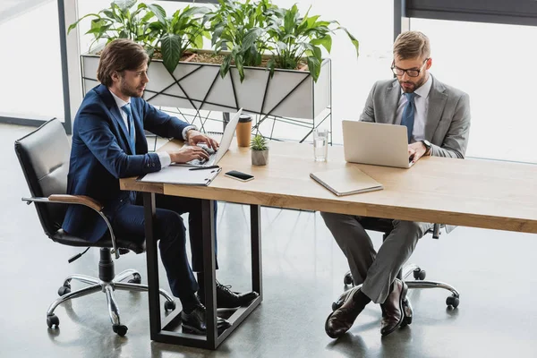 Vista de ángulo alto de los jóvenes hombres de negocios en trajes usando computadoras portátiles y trabajando juntos en la oficina - foto de stock