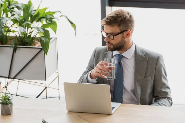 Jeune homme d'affaires tenant un verre d'eau et regardant loin tout en utilisant un ordinateur portable dans le bureau — Photo de stock