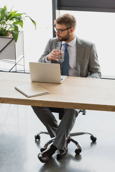 Handsome young businessman in eyeglasses holding glass of water and using laptop — Stock Photo