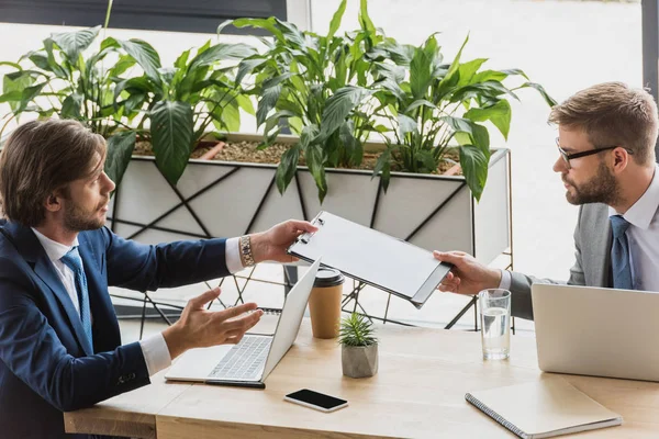 Jóvenes empresarios que sostienen el portapapeles y se miran mientras trabajan con computadoras portátiles en la oficina - foto de stock