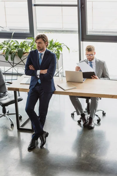 High angle view of young businessman with crossed arms looking away while colleague holding clipboard and using laptop behind — Stock Photo