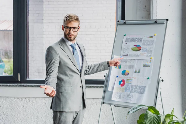 Handsome young businessman in suit and eyeglasses pointing at whiteboard with charts and looking at camera — Stock Photo