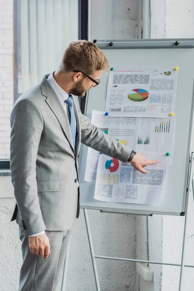 Handsome young businessman in suit and eyeglasses pointing at whiteboard with charts and graphs — Stock Photo