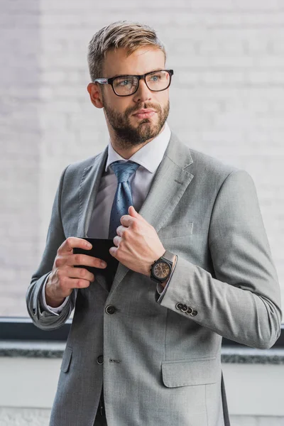 Handsome young businessman in suit and eyeglasses holding smartphone and looking away — Stock Photo