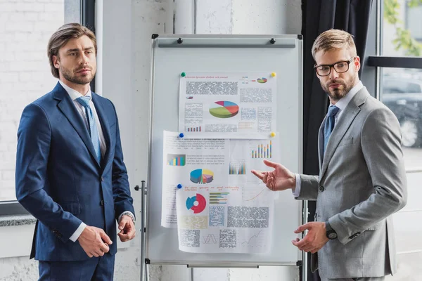 Jóvenes hombres de negocios guapos de pie cerca de pizarra blanca con gráficos y gráficos - foto de stock