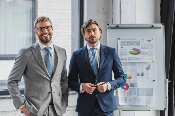 Trois beaux jeunes hommes d'affaires en costumes debout ensemble et regardant la caméra dans le bureau — Photo de stock