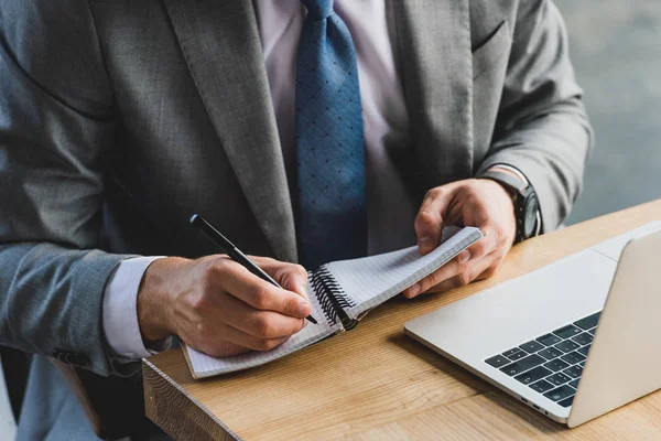 Cropped shot of businessman in formal wear taking notes in notebook and using laptop at workplace — Stock Photo