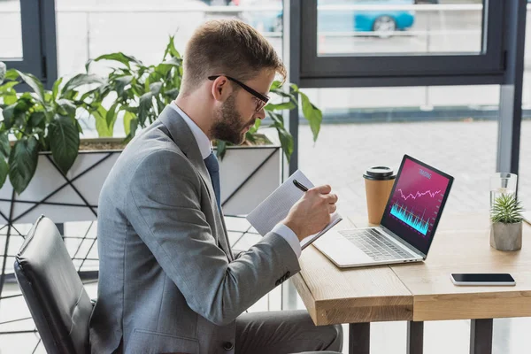 Hombre de negocios con traje y anteojos tomando notas en cuaderno y utilizando portátil con gráficos y gráficos de negocios — Stock Photo