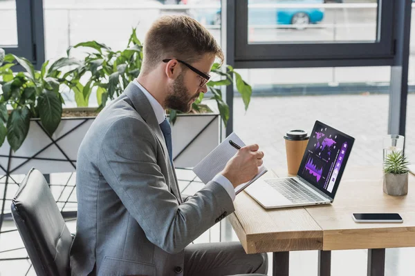 Joven hombre de negocios en traje y anteojos tomando notas en el cuaderno y el uso de ordenador portátil con gráficos de negocios - foto de stock