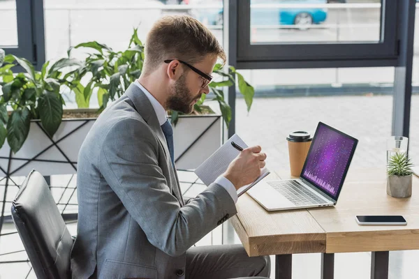 Young businessman in suit and eyeglasses taking notes in notebook and using laptop with business graphs — Stock Photo