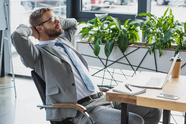 Young businessman in suit and eyeglasses sitting with hands behind head in office — Stock Photo