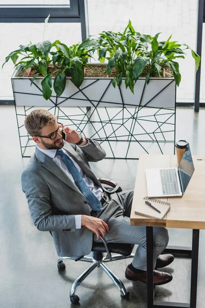 Vista de ángulo alto del hombre de negocios sonriente hablando por teléfono inteligente y utilizando el ordenador portátil en la oficina - foto de stock